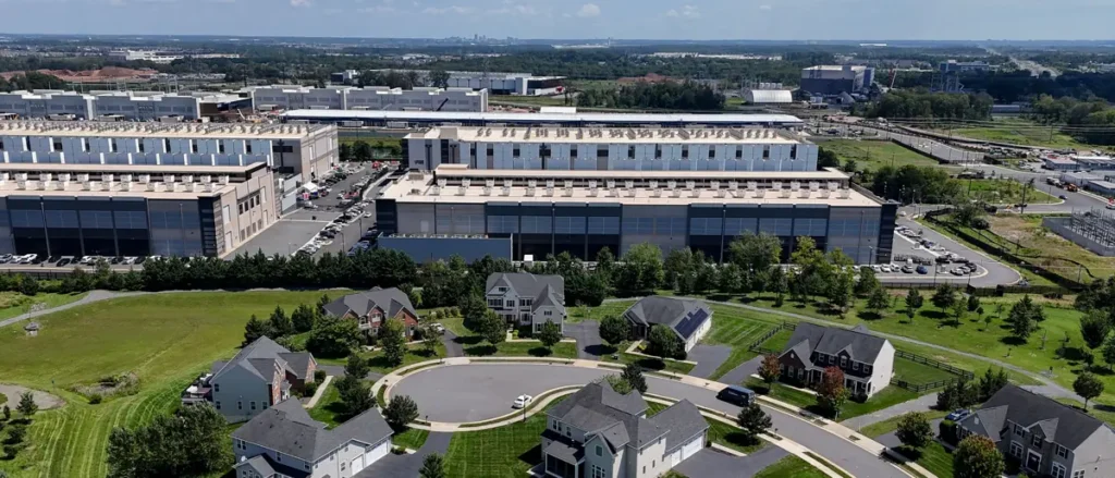 The image shows an aerial view of a suburban neighborhood with residential houses surrounded by greenery, adjacent to a large industrial complex with multiple buildings and parking lots.