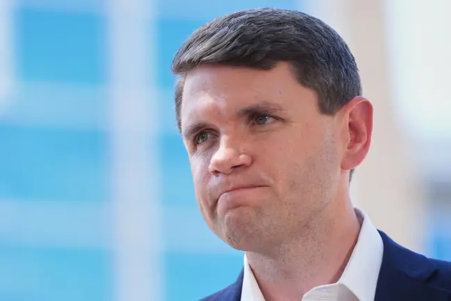 The image shows a man in a suit with a serious expression, set against a blurred background with blue and white tones.