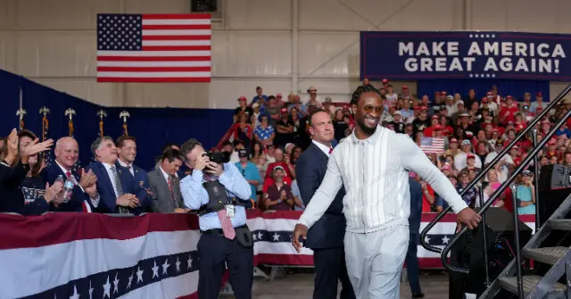 A man in a white outfit walks down a stage at a rally with a crowd and an "MAKE AMERICA GREAT AGAIN!" sign in the background.