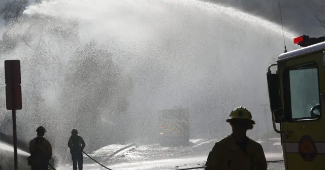 Firefighters are seen managing a large water spray from a fire truck, likely as part of a firefighting operation.
