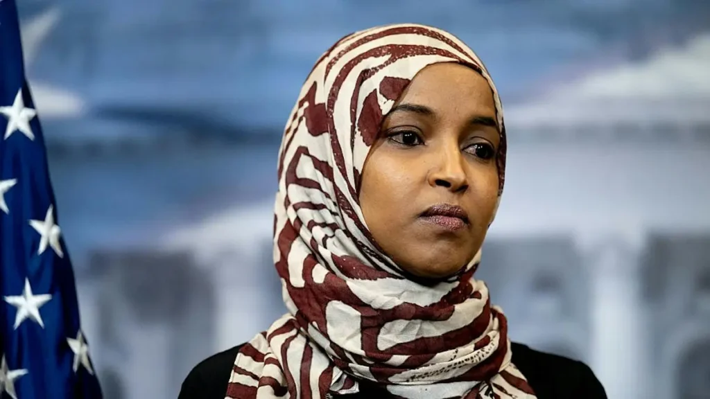 The image shows a person wearing a patterned hijab, standing in front of an American flag, suggesting a political or public setting.
