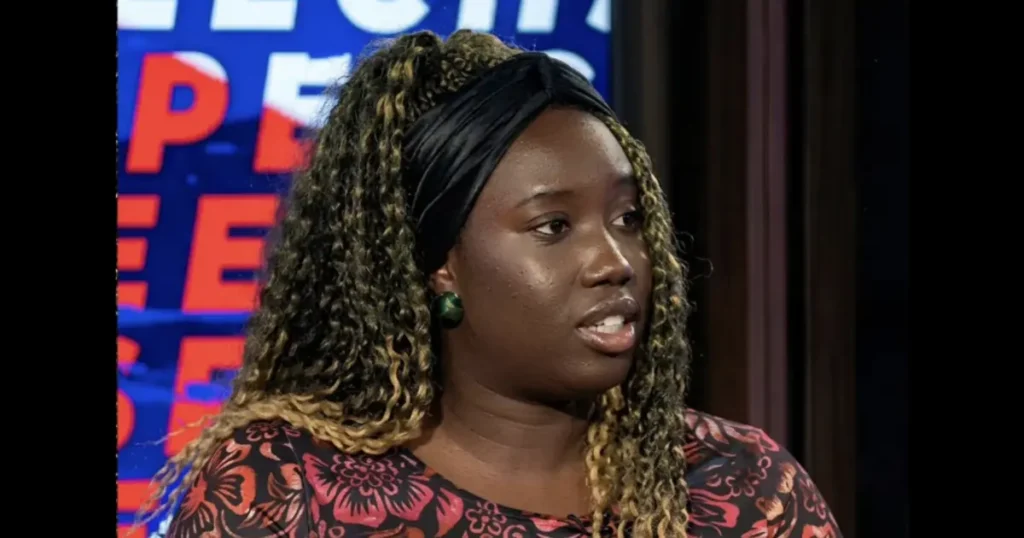 The image shows a woman with curly hair wearing a floral top and a headband, speaking in what appears to be a news studio setting.