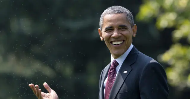The image shows a person in a suit and tie, smiling and gesturing with their hand, with a blurred background of trees and foliage.