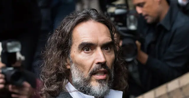 The image shows a man with long hair and a beard, surrounded by photographers and cameras, suggesting a public or media event.
