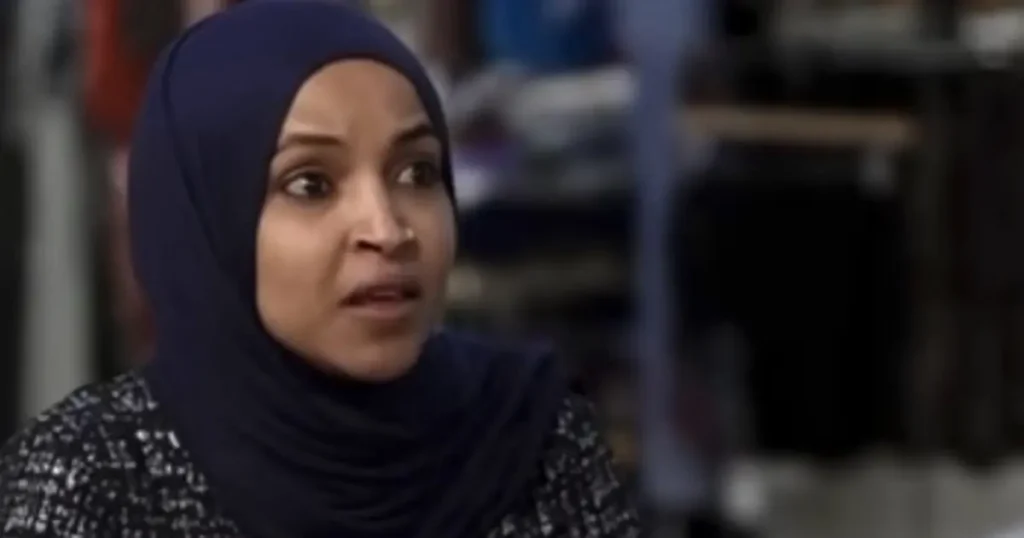 The image shows a person wearing a dark blue hijab, seated in what appears to be an indoor setting with blurred background elements.