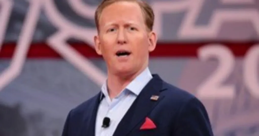 The image shows a man in a suit speaking at a podium, with a backdrop featuring a logo and the year "2020."