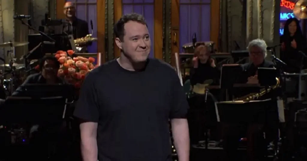 The image shows a man in a black shirt standing in front of a band and a floral arrangement, likely during a performance or event.