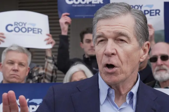 A man in a suit is speaking at a political rally, with supporters holding signs that read "ROY COOPER."