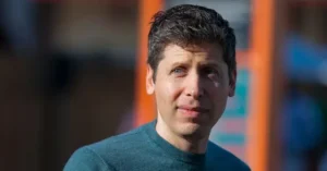 The image shows a person wearing a teal sweater, standing outdoors with a blurred background featuring orange and blue elements.