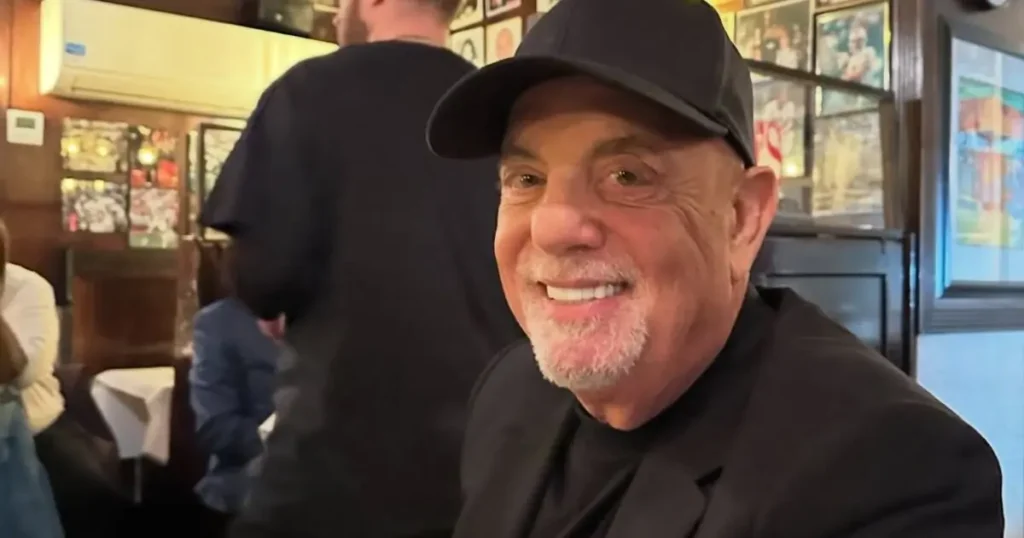The image shows a man smiling and wearing a black cap and suit, seated in a restaurant with framed pictures on the wall in the background.