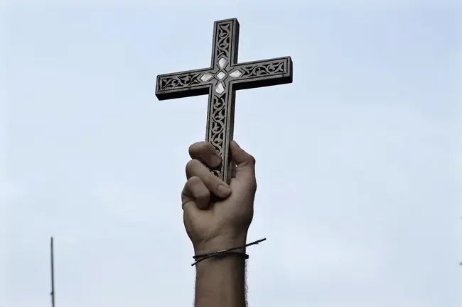 A hand holds a cross against a cloudy sky, symbolizing faith or a religious gesture.