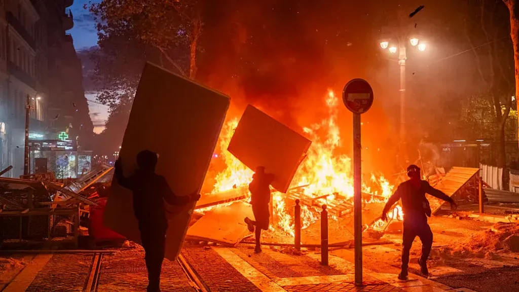 The image depicts a nighttime scene of a protest or riot, with individuals carrying large objects and a large fire burning in the background.