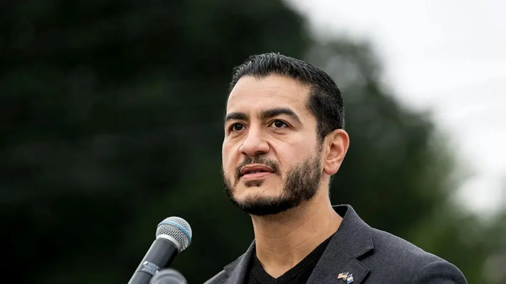 The image shows a man speaking into a microphone, dressed in a suit, with a pin on his lapel, set against a backdrop of trees and an overcast sky.