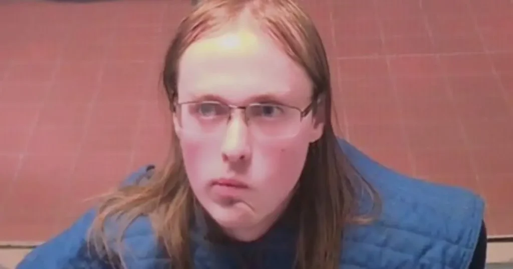 The image shows a person with long hair and glasses, wearing a blue vest, against a brick wall background.