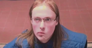 The image shows a person with long hair and glasses, wearing a blue vest, against a brick wall background.
