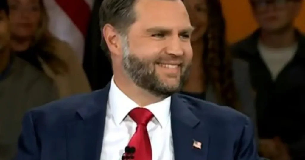 The image shows a man in a suit and red tie, smiling, with an American flag pin on his lapel, in what appears to be a formal setting.