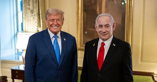 The photo shows two individuals in formal attire, smiling and standing side by side in a well-lit room with elegant decor.