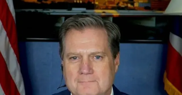 The image shows a man in a suit with an American flag and another flag in the background, suggesting a formal or official setting.