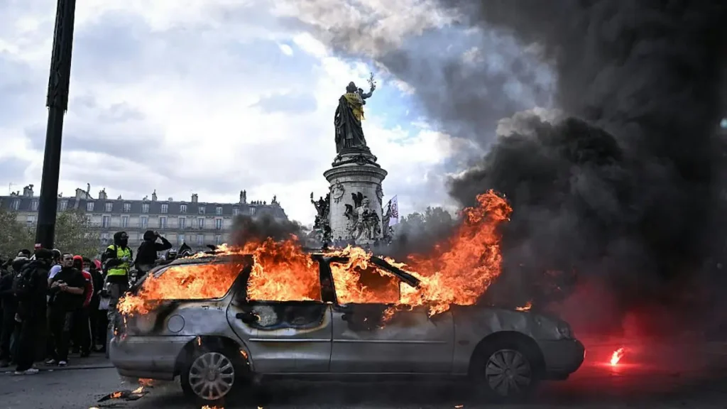 A car is engulfed in flames during a protest, with smoke and debris filling the air, as people gather around in the background.