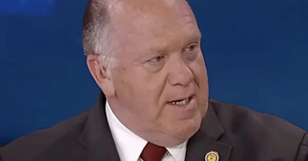 The image shows a man in a suit and tie, speaking in what appears to be a formal or professional setting.