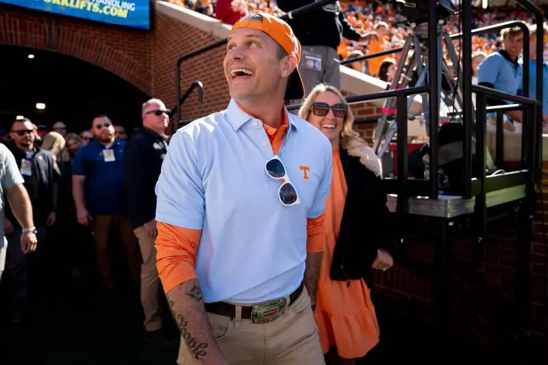A man in a light blue polo shirt and orange cap is smiling and walking through a crowd at a sports event.