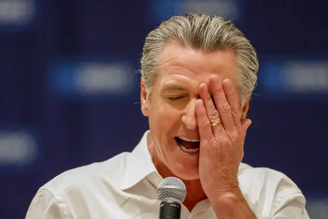 A man in a white shirt is speaking into a microphone while covering his face with his hand, possibly indicating frustration or exhaustion.