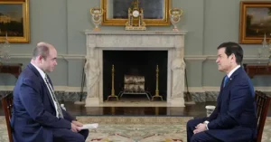 Two men in suits are seated in a formal setting, engaged in a conversation in front of an ornate fireplace.