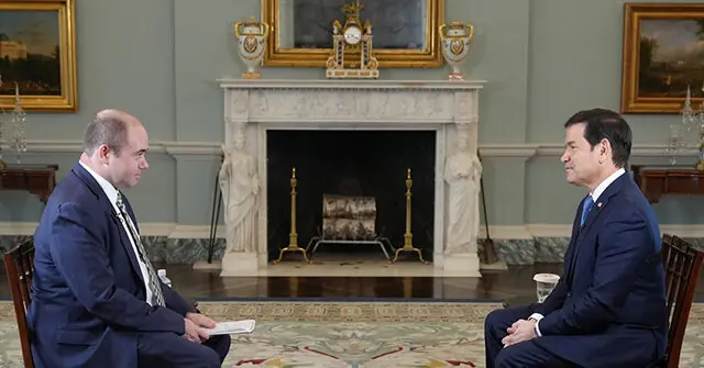 Two men in suits are seated in a formal setting, engaged in a conversation in front of an ornate fireplace.