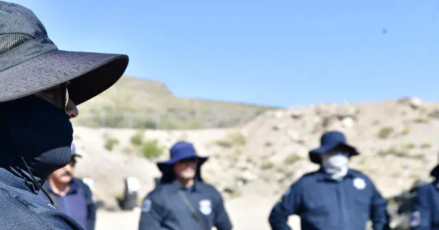 A group of individuals wearing hats and uniforms stands in a desert-like environment under a clear blue sky.