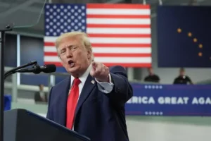 The image shows a political figure giving a speech at a podium with an American flag and a sign reading "Make America Great Again" in the background.