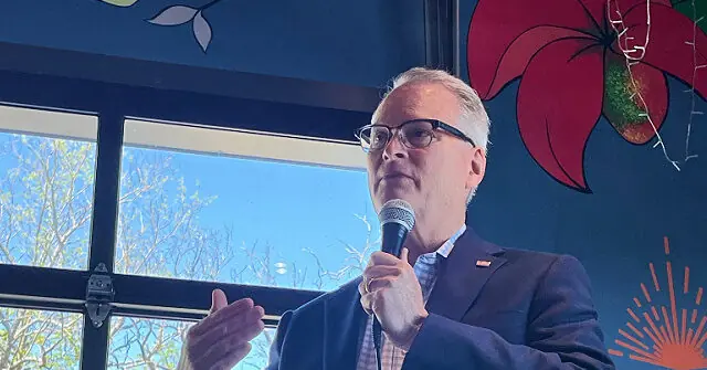 A man in a suit is speaking into a microphone, gesturing with his hand, in front of a colorful mural and a window with a view of trees.