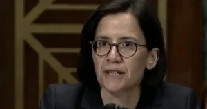 The image shows a person wearing glasses and a suit, speaking in what appears to be a formal setting.