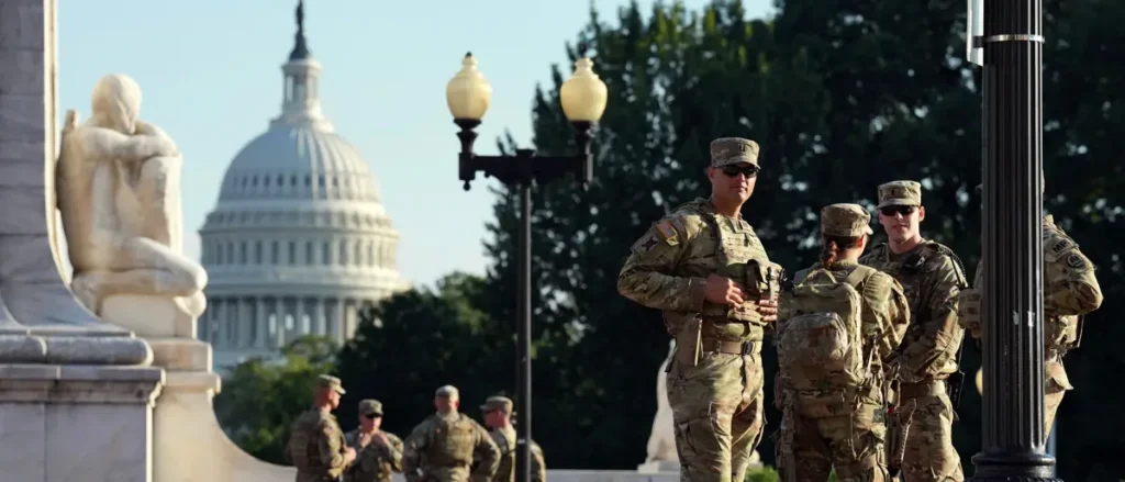 A group of soldiers in camouflage uniforms stands near a lamp post with the U.S. Capitol building in the background.