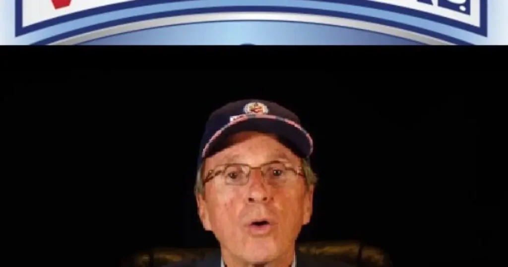 The image shows a man wearing a cap and glasses, speaking in front of a backdrop with a logo.