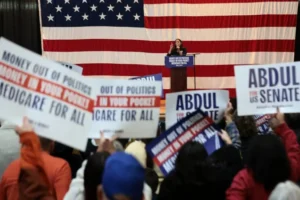 A political rally is taking place with a speaker at a podium and supporters holding signs advocating for "Medicare for All" and "Money Out of Politics."