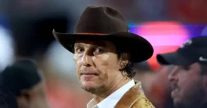 The image shows a person wearing a cowboy hat and a white shirt, with a blurred background suggesting a public or event setting.