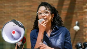 A woman is speaking into a microphone while holding a megaphone, suggesting she is addressing a crowd or leading a protest.
