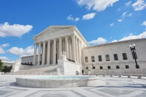The image shows the Supreme Court Building in Washington, D.C., under a bright blue sky with scattered clouds.