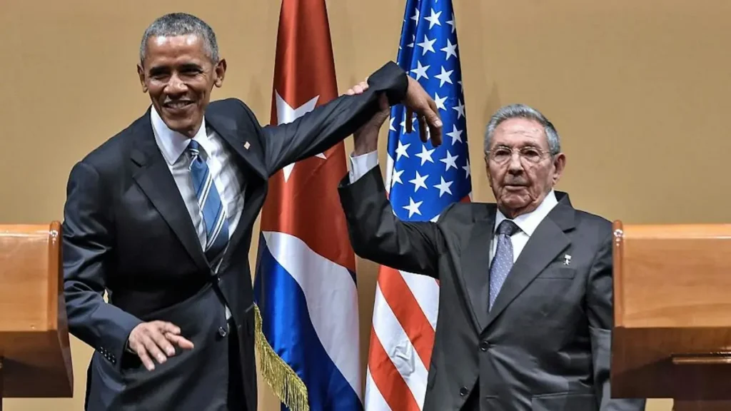 The photo shows two leaders in formal attire shaking hands in front of the flags of the United States and Cuba, symbolizing a diplomatic gesture.