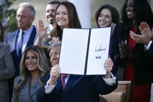 The image shows a person holding up a signed document in front of a group of people who are clapping and smiling.