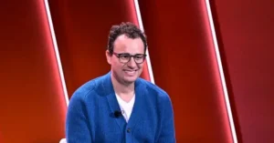The image shows a person wearing a blue cardigan and glasses, sitting in front of a red and white backdrop, smiling.