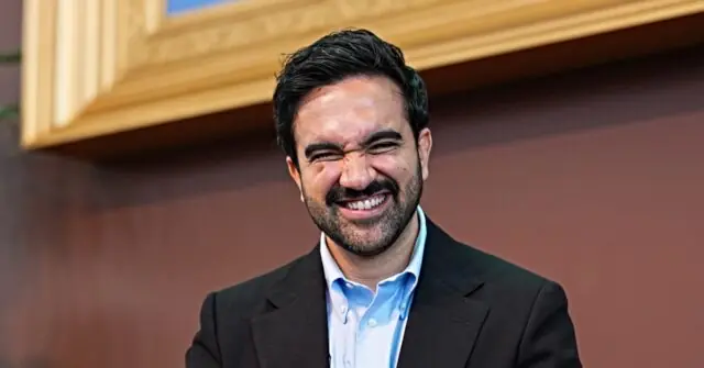 The image shows a man in a suit smiling warmly, with a background that includes wooden cabinets.