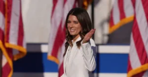 A woman in a white outfit is smiling and clapping in front of American flags.