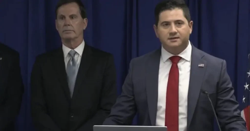 Two men in suits stand at a podium with an American flag in the background, suggesting a formal press conference or official announcement.