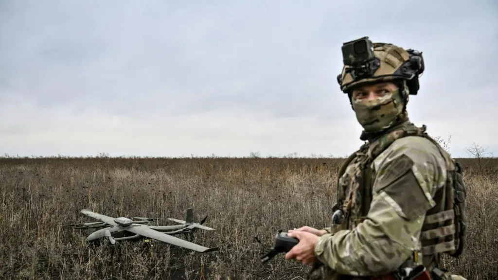 A soldier operates a drone in a field, equipped with tactical gear and a camera mounted on their helmet.