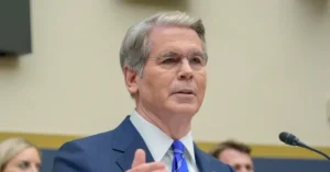 The image shows a man in a suit and tie speaking at a formal event, with a microphone in front of him.