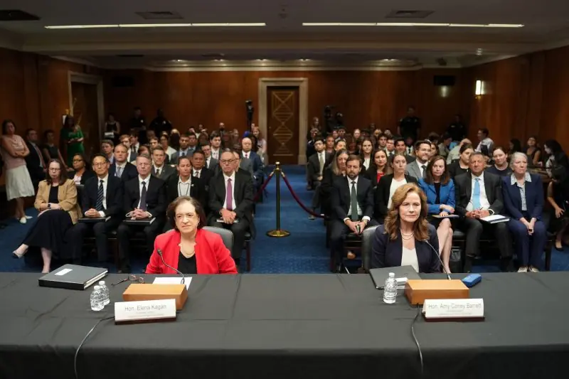 The image depicts a formal hearing or meeting with two individuals seated at a table, facing an audience in a large room.