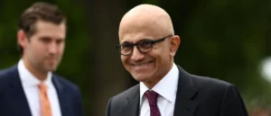 The image shows a man in a suit and tie smiling, with another person partially visible in the background.