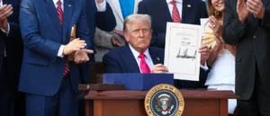 The image shows a formal event where a person is holding a document and standing at a podium with the seal of the President of the United States, while others clap and observe.