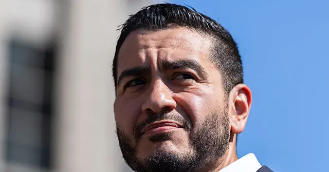 The image shows a close-up of a man with a beard and short hair, wearing a white shirt, against a backdrop of a clear blue sky and a building.